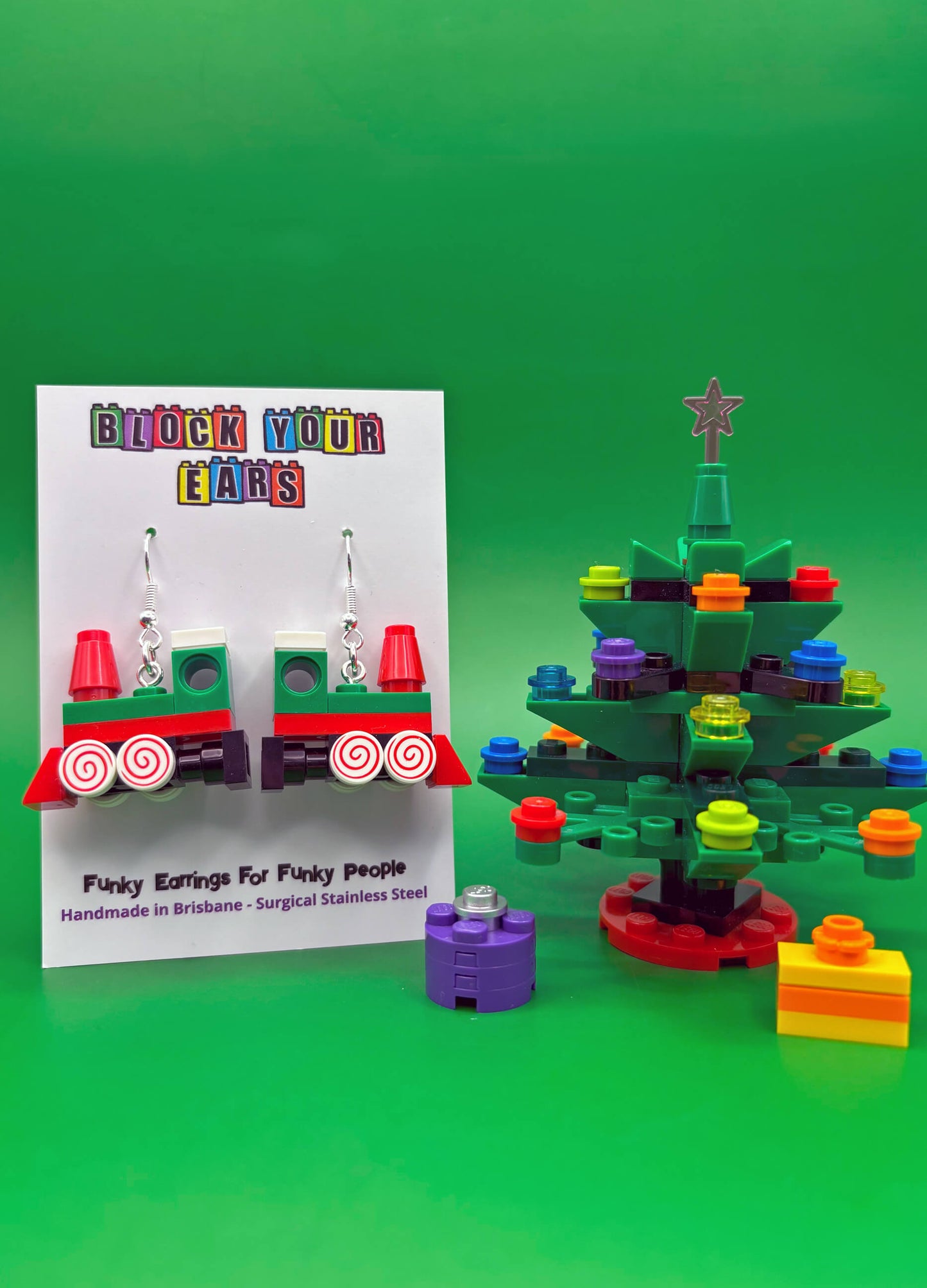 Colorful train earrings on a display card with a LEGO-themed Christmas tree on a green background