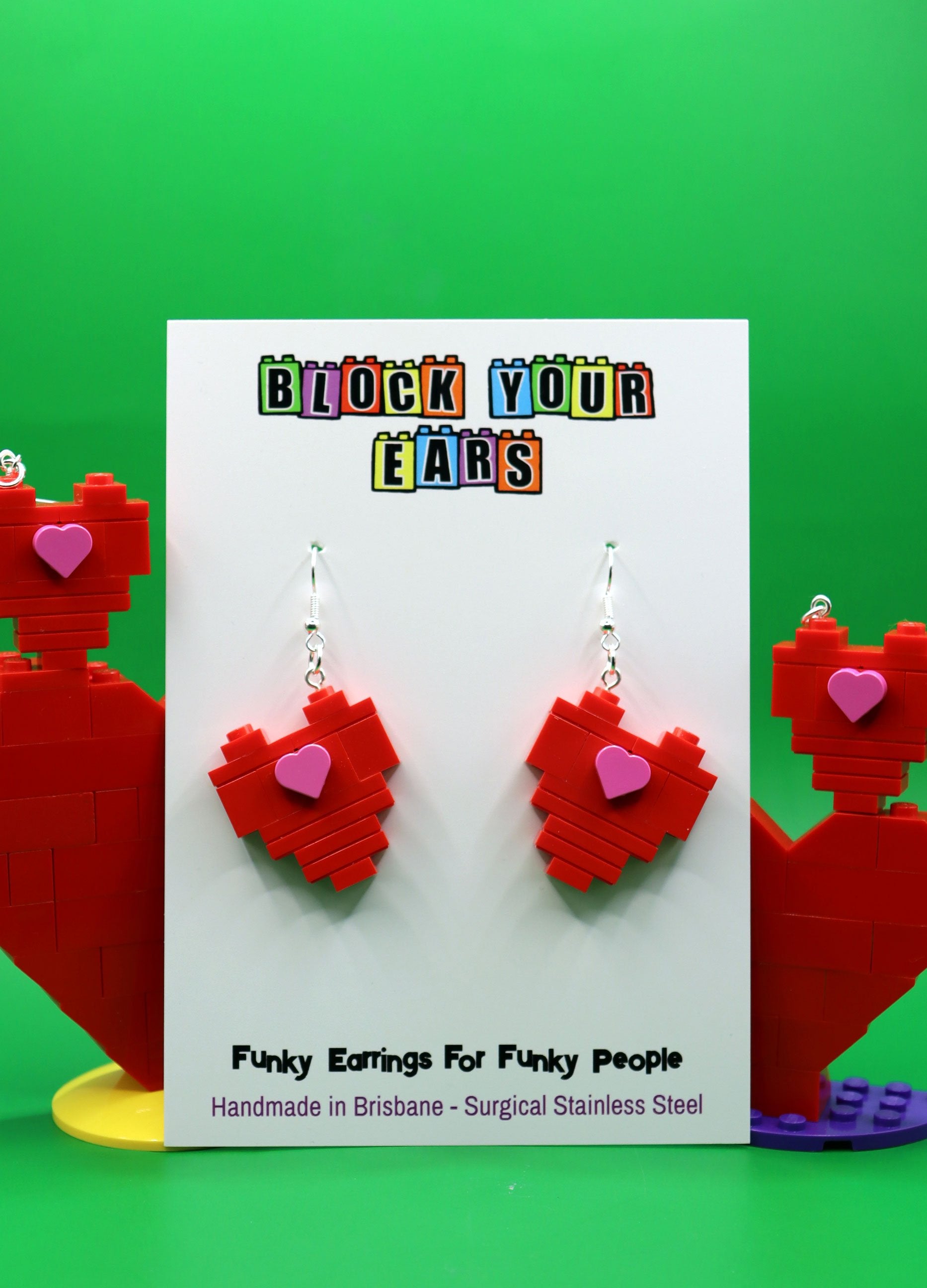 Red heart-shaped LEGO earrings on a 'Block Your Ears' branded card with a green background