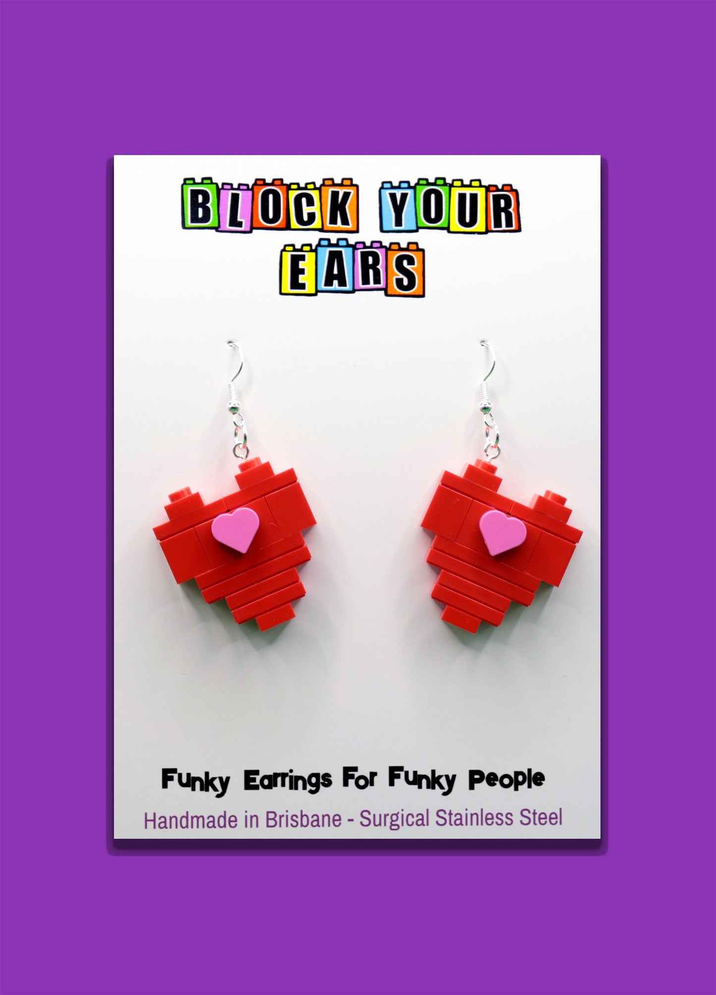 Red heart-shaped LEGO earrings on a card with 'Block Your Ears' branding on a purple background